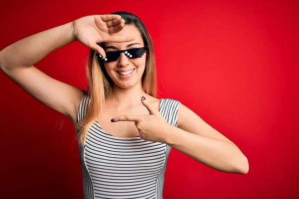 Young beautiful brunette woman wearing funny thug life sunglasses over red background smiling making frame with hands and fingers with happy face. Creativity and photography concept.
