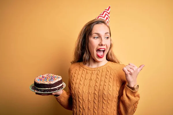 Young beautiful blonde woman holding birthday cake over isolated yellow background pointing and showing with thumb up to the side with happy face smiling