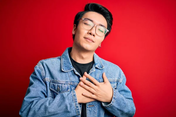 Young handsome chinese man wearing denim jacket and glasses over red background smiling with hands on chest with closed eyes and grateful gesture on face. Health concept.