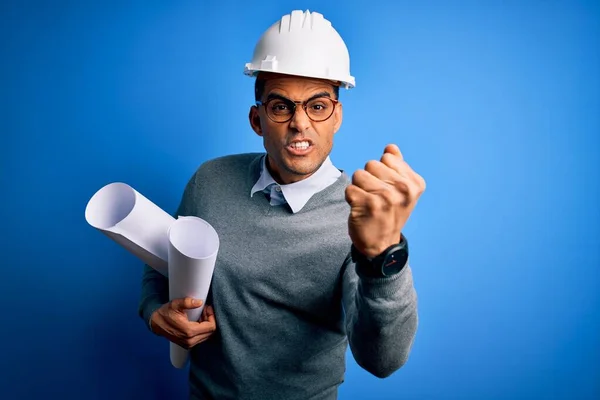 Young african american architect man wearing safety helmet holding bluprints annoyed and frustrated shouting with anger, crazy and yelling with raised hand, anger concept