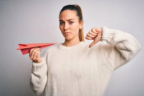 Beautiful blonde woman with blue eyes holding red paper airplane over white background with angry face, negative sign showing dislike with thumbs down, rejection concept