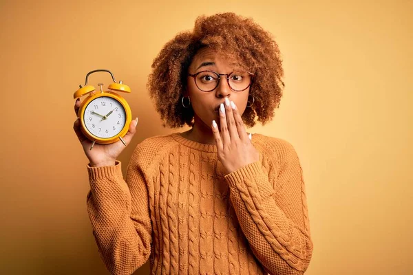 Young beautiful African American afro woman with curly hair holding vintage alarm clock cover mouth with hand shocked with shame for mistake, expression of fear, scared in silence, secret concept