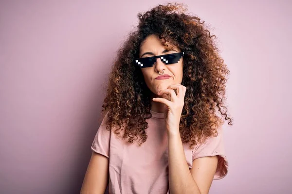 Young beautiful woman with curly hair and piercing wearing funny thug life sunglasses with hand on chin thinking about question, pensive expression. Smiling with thoughtful face. Doubt concept.