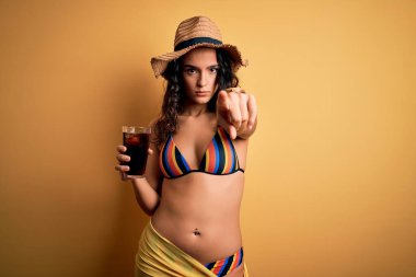 Genç, kıvırcık saçlı, tatilde bikini giyen, kola içen, kamerayı işaret eden, el işareti yapan, ön taraftan olumlu ve kendinden emin bir jest yapan güzel bir kadın.