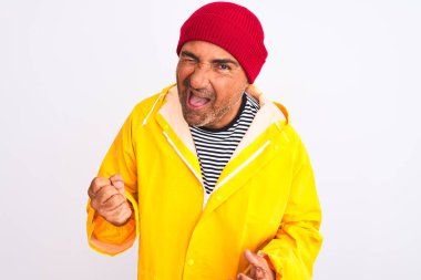 Yağmurluk ve yünlü şapka giyen orta yaşlı bir adam izole edilmiş beyaz arka planda dikilip kameraya seksi bir ifadeyle bakıyor, neşeli ve mutlu bir yüzle..