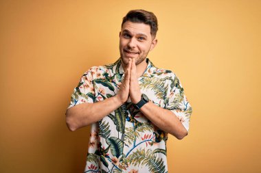 Tatilde mavi gözlü, çiçekli bir tişört giyen, sarı arka planda dua eden ellerini birleştirip af dileyen, kendinden emin gülümseyen genç bir adam..