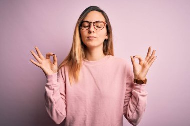 Mavi gözlü, pembe arka planda süveter ve gözlük takan güzel sarışın kadın rahatla ve parmaklarıyla meditasyon hareketi yaparken gözleri kapalı gülümse. Yoga konsepti.