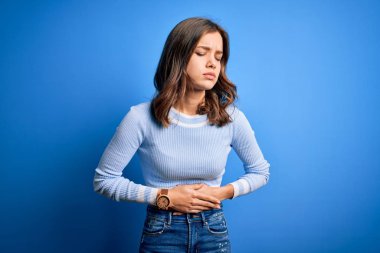 Mavi izole edilmiş arka plan üzerinde duran sıradan bir kazak giyen genç, güzel sarışın kız midesi üzerinde el ele tutuşuyor çünkü hazımsızlık, acı verici bir hastalık kendini iyi hissetmiyor. Ache kavramı.