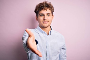 Sarışın, kıvırcık saçlı, beyaz arka planda çizgili tişört giyen, neşeli, yardım eli uzatan ve kabul gören genç bir adam..