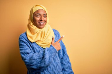 Genç Afrikalı Afro kadın, izole edilmiş sarı arka planda tesettürlü, neşeli, yüzünde mutlu ve doğal bir ifadeyle, elleri ve parmaklarıyla yüzünü gösteren bir tebessüm takıyor.
