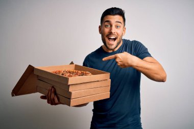 Beyaz zemin üzerinde İtalyan pizzası olan teslimat kutularını tutan yakışıklı genç adam mutlu bir şekilde elini ve parmağını gösterdi.