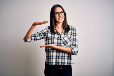 Mavi gözlü genç esmer kadın, sıradan gömlek ve gözlük takıyor beyaz arka planda, elleri büyük ve büyük beden işareti, ölçü sembolü gösteren hareketler yapıyor. Kameraya bakıp gülümsüyor. Ölçüm konsepti.