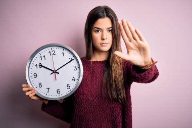 Geri sayım yapan güzel bir kız pembe arka planda büyük bir saat tutuyor. Açık elle dur işareti yapıyor. Ciddi ve kendinden emin bir ifadeyle, savunma hareketi yapıyor.