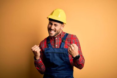 Sarı arka plan üzerinde inşaat üniforması ve güvenlik kaskı takan genç müteahhit kollarını kaldırıp, gülümseyerek ve başarı çığlıkları atarak çok mutlu ve heyecanlı bir jest yapıyor. Kutlama konsepti.
