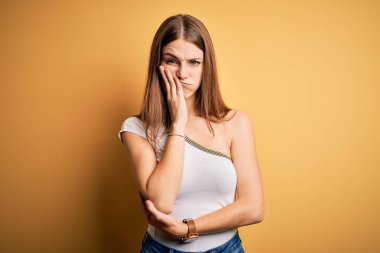 Genç, güzel, kızıl saçlı bir kadın sarı arka plan üzerine günlük tişört giyip kollarını kavuşturarak depresyondan sıkılmış ve yorgun görünüyor..