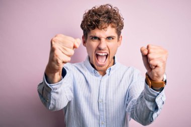 Sarışın, kıvırcık saçlı, beyaz arka planda çizgili gömlek giyen kızgın ve öfkeli genç bir adam öfkeyle bağırırken yumruklarını kaldırıyor. Öfke ve agresif kavram.