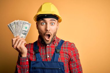 Güvenlik kaskı takan genç müteahhit elinde dolar tutarken sarı arka planda şok geçirmiş, korkmuş ve korku ifadesinden heyecanlanmış.