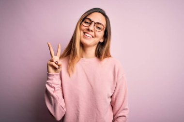Mavi gözlü güzel sarışın kadın pembe arka planda süveter ve gözlük takıyor. İki numaralı parmaklarıyla işaret ediyor ve gülümsüyor. Kendinden emin ve mutlu..