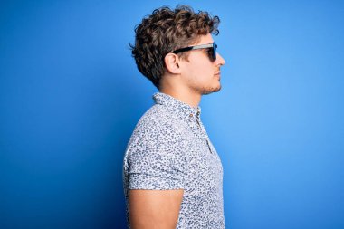 Genç, sarışın, kıvırcık saçlı, tatilde çizgili polo ve güneş gözlüğü takan, kendine güveni tam, rahat bir poz veren yakışıklı bir adam..