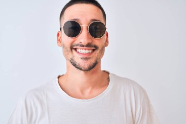 Güneş gözlüğü takan genç yakışıklı bir adam ve izole edilmiş arka planda rahat bir tişört giyen mutlu bir yüzü olan ve kendine güvenen bir gülümsemeyle dişlerini gösteren.