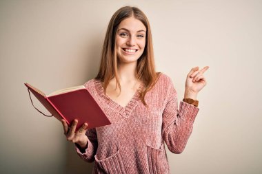Genç, güzel, kızıl saçlı, kitap okuyan kız izole edilmiş beyaz arka plan üzerinde çok mutlu bir şekilde el ve parmak uçlarını işaret ediyor.