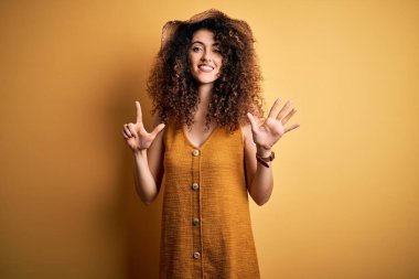 Güzel esmer kadın tatilde. Kıvırcık saçlı, piercing takan, şapka takan, elbise giyen yedi numaralı parmaklarıyla işaret eden mutlu ve kendinden emin bir şekilde gülümseyen..