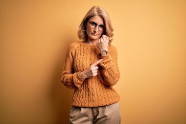 Sarışın, orta yaşlı bir kadın sarı arka planda gözlüklü, sıradan bir süveter giyiyor. Saati, sabırsızlığı ve rahat bir ifadeyle kameraya bakıyor.