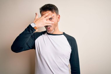 Genç ve yakışıklı bir adam, günlük tişört giyip izole edilmiş beyaz arka planda dikiliyor. Şok olmuş bir şekilde yüzünü ve gözlerini kapıyor. Utanmış bir ifadeyle parmaklarının arasından bakıyor..