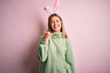 Paskalya tavşanı kulakları takan genç ve güzel bir kadın izole edilmiş pembe arka planda durup gülümsüyor telefonla el ve parmak hareketleriyle telefonla konuşuyor gibi. İletişim kavramları.