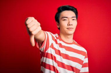 Kırmızı arka planda dikilen sıradan çizgili tişört giyen genç, yakışıklı Çinli bir adam mutsuz, reddedilmiş ve olumsuz hareketlerde bulunmuş. Kötü ifade.