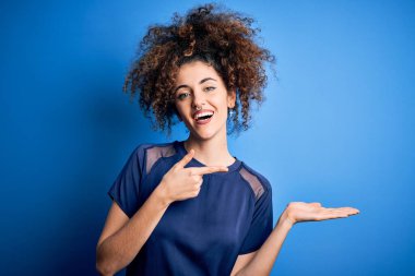 Kıvırcık saçlı ve piercingli genç bir kadın. Sıradan mavi tişörtler giyiyor. Kameraya hayret ediyor ve gülümsüyor..