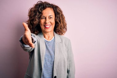 Soyutlanmış pembe arka planda zarif bir ceket giyen, orta yaşlı, güzel bir iş kadını selamlama ve hoş geldin demek için gülümseyerek el sıkışır. Başarılı bir iş..