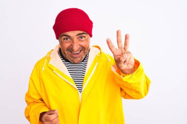 Yağmurluk ve yün şapka giyen orta yaşlı bir adam izole edilmiş beyaz arka plan üzerinde duruyor ve üç numaralı parmaklarıyla işaret ederken kendinden emin ve mutlu bir şekilde gülümsüyor..