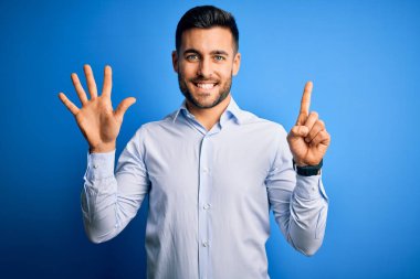 Zarif gömlekli genç adam mavi arka planda dikiliyor ve 6 numaralı parmaklarıyla işaret ederken kendinden emin ve mutlu bir şekilde gülümsüyor..