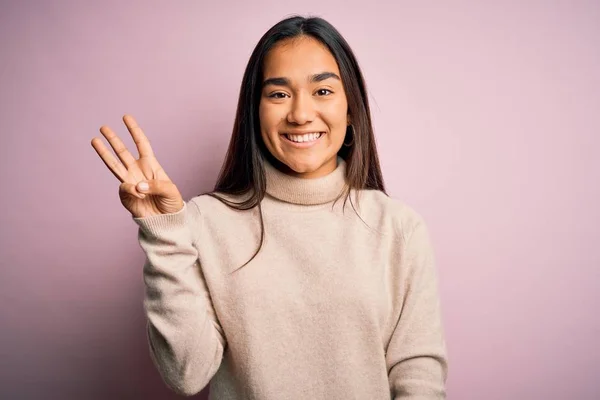 若いです美しいですアジアの女性身に着けていますカジュアルTurtleeckセーター上のピンクの背景表示と指差しますと指番号3とともに笑顔自信と幸せ — ストック写真