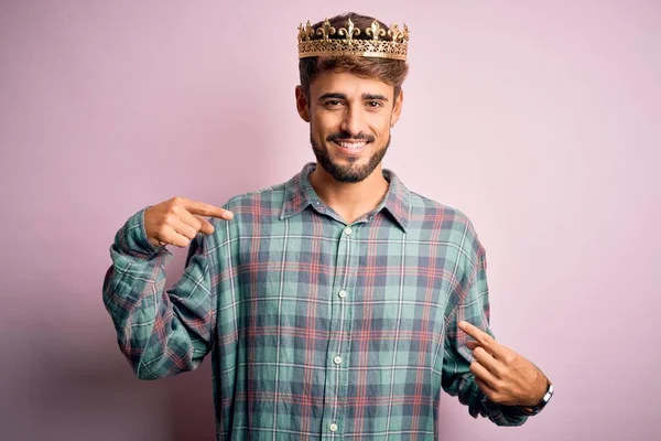 Young man with beard wearing golden crown of king standing over isolated pink background looking confident with smile on face, pointing oneself with fingers proud and happy.