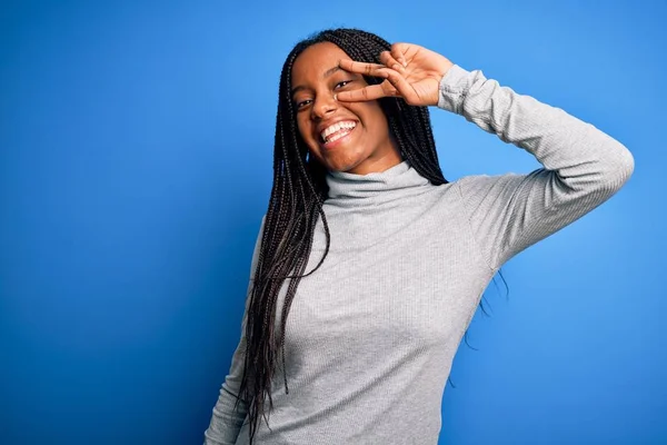 Young african american woman standing wearing casual turtleneck over blue isolated background Doing peace symbol with fingers over face, smiling cheerful showing victory