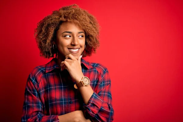 Young beautiful African American afro woman with curly hair wearing casual shirt with hand on chin thinking about question, pensive expression. Smiling with thoughtful face. Doubt concept.