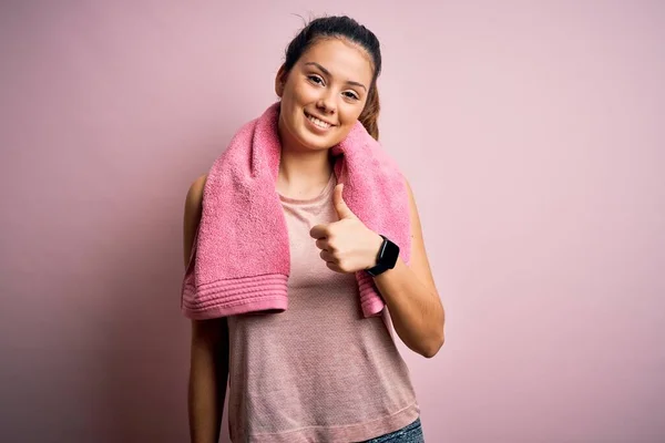 ピンクの背景にスポーツウェアとタオルを身に着けている若い美しいブルネットのスポーツウーマンは 手で幸せな親指を上げるジェスチャーをしています 成功を示すカメラを見て表現を承認する — ストック写真