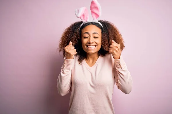 年轻的非洲裔美国女人 有着一头浓密的黑发 粉红的背景上长着小兔子般的耳朵 兴奋地期待着成功 举起双臂 紧闭双眼笑着庆祝胜利 赢的概念 — 图库照片