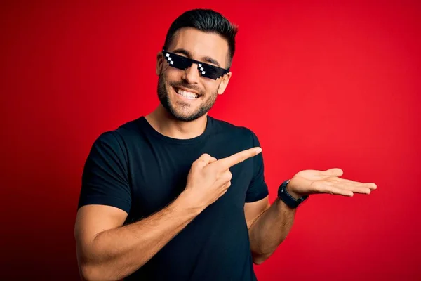 Young handsome man wearing funny thug life sunglasses over isolated red background amazed and smiling to the camera while presenting with hand and pointing with finger.