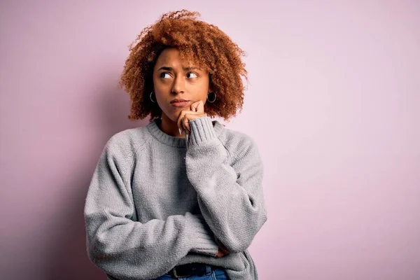 Young beautiful African American afro woman with curly hair wearing casual sweater with hand on chin thinking about question, pensive expression. Smiling with thoughtful face. Doubt concept.