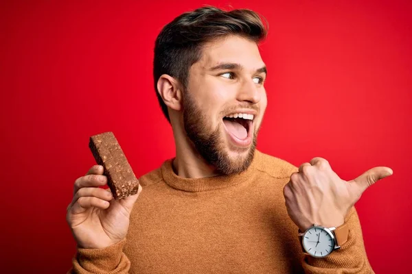 年轻的金发男子 留着胡子 蓝眼睛 正吃着健康的巧克力蛋白质棒 指着大拇指朝外看去 笑容满面 — 图库照片