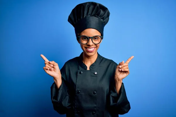 年轻的非洲女厨师身穿炊具制服 头戴蓝色背景的帽子 带着自信的微笑 手指指向不同的方向 广告的复制空间 — 图库照片