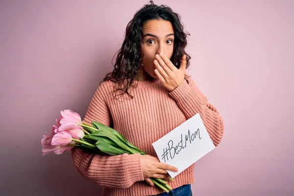 Beautiful curly hair woman holding best mom message and tulips celebrating mothers day cover mouth with hand shocked with shame for mistake, expression of fear, scared in silence, secret concept