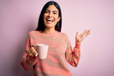 Genç ve güzel İspanyol kadın pembe izole edilmiş arka planda bir fincan sıcak kahve içiyor mutlu ve heyecanlı, kazanan yüz ifadesi zaferi büyük bir gülümsemeyle kutluyor ve ellerini kaldırıyor.