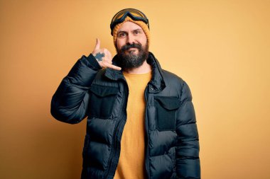 Sakallı, kar maskesi takmış, kar maskesi takmış ve kayak gözlüklü yakışıklı kayakçı telefonla el ve parmaklarıyla telefonla konuşuyor gibi gülümsüyor. İletişim kavramları.