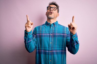 Gündelik gömlek ve gözlük takan genç yakışıklı adam pembe arka planda durmuş şaşkın şaşkın yukarı bakıyor ve parmaklarıyla işaret ediyor ve kollarını kaldırıyordu..