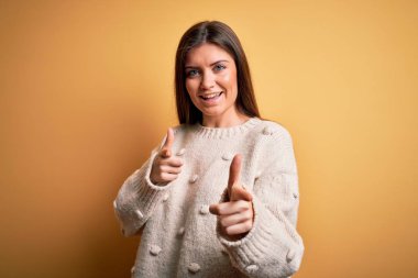 Sarı arka planda dikilip mutlu ve komik yüzlü parmakları kameraya gösteren mavi gözlü genç ve güzel bir kadın. İyi enerji ve titreşimler.