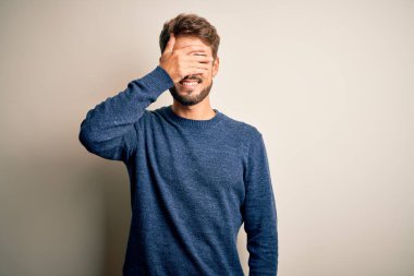 Sakallı, sıradan bir süveter giyen, beyaz arka planda dikilen, gülümseyen ve yüzünde şaşkınlıktan gözleri kapayan genç yakışıklı bir adam. Kör kavram.